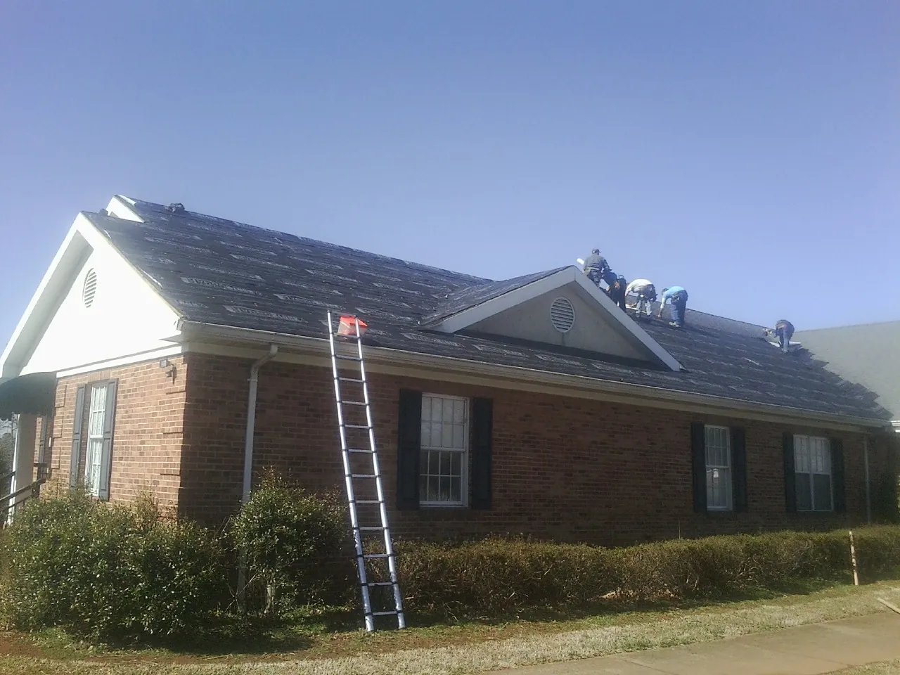 Crew tearing shingles off a brick ranch roof with a ladder set — Metal Roof Repair work by our crew in Fort Shaw