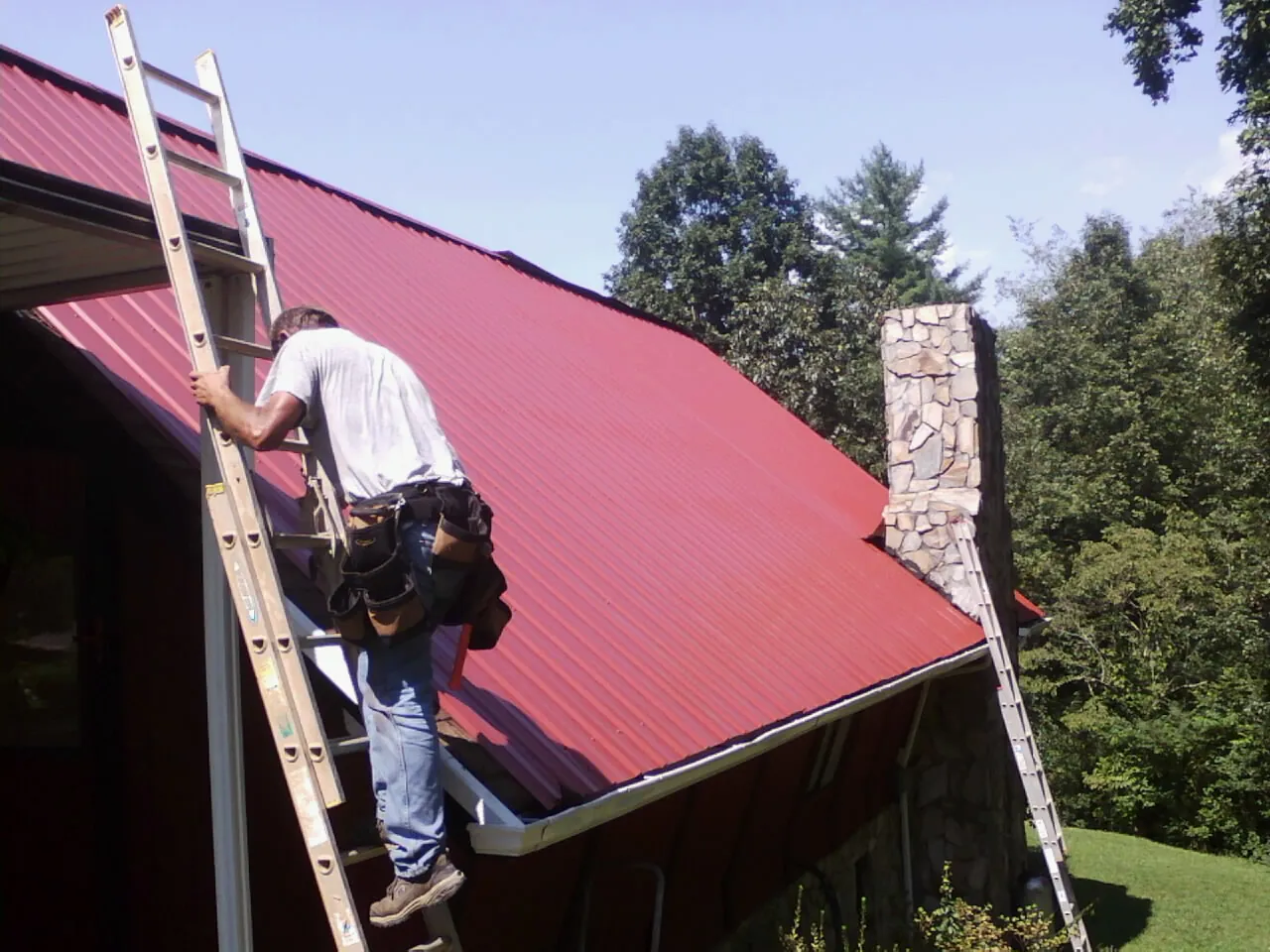Roofer on a ladder beside a red metal roof and stone chimney — Soffit & Fascia Repair by Roofing Brothers in Fort Shaw